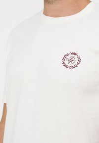 Κοντινό πλάνο ενός ατόμου που φοράει λευκό T-shirt με ένα μικρό μπορντό έμβλημα με κορώνα και στεφάνι δάφνης στο στήθος.