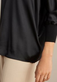 Top lungo a maniche nere in satin con una texture liscia e una vestibilità comoda, caratterizzato da polsini aderenti e cuciture sottili, abbinato a pantaloni beige chiaro.