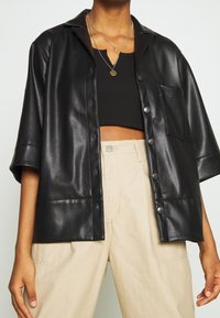 Personne portant un crop top noir, une chemise en cuir noire à manches courtes et boutons, un pantalon beige taille haute et des colliers superposés.