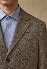 Brown herringbone-patterned blazer made of textured wool, featuring notched lapels, a chest pocket, and a button closure, layered over a light blue shirt.