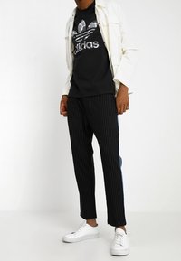 Muž oblečený v černém tričku s kamuflážním logem Adidas, krémovém kabátu, černých kalhotách s pruhy a modrým detailem, a bílých teniskách na bílém pozadí.