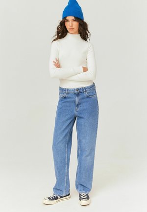 Haut long à manches blanches côtelé, jean bleu en denim ample et baskets marines. Le mannequin porte un bonnet tricoté bleu vif; fond neutre.