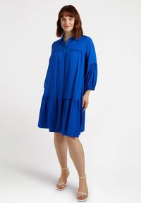 Abito camicia blu realizzato in un tessuto leggero, con una chiusura frontale a bottoni, maniche a sbuffo e orlo a balze. Abbinato a sandali con cinturini.
