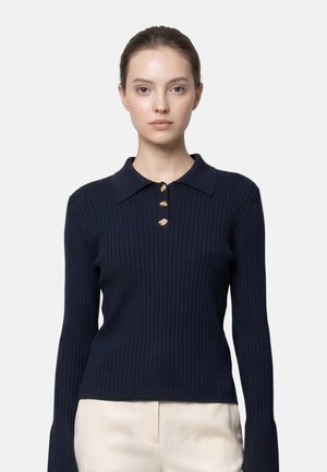 Frau mit zurückgebundenem Haar trägt ein marineblau geripptes Langarm-Poloshirt mit drei goldenen Knöpfen und hellen Hosen.