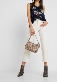 Haut floral marine, pantalon beige clair et bottines à imprimé serpent grises. Le sac est un petit sac à main avec un motif serpent similaire.