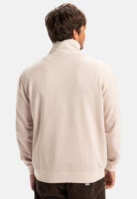 Light pink sweatshirt with a high collar, soft texture, ribbed cuffs, and a logo detail on the left sleeve. Worn with dark brown pants.