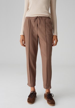 Bruine, op maat gemaakte broek met een taillekoord, voorzien van plooien aan de voorkant en taps toelopende pijpen. Gecombineerd met bruine suède sneakers.