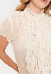 Blouse légère en beige transparent avec des détails froncés le long du col et des boutons, présentant un tissu texturé et fluide avec des accents verts subtils.