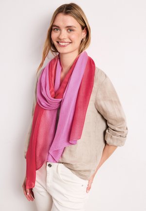 Hymyilevä nainen, jolla on vaaleat hiukset, pukeutuu beigeen paitaan ja suurikokoiseen vaaleanpunaiseen ja punaiseen huiviin, seisoo valkoiset housut taskussa yhdellä kädellä.