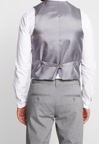 Chaleco de satén gris con una textura suave, que cuenta con una correa trasera y detalle de hebilla. Lleva puesto sobre una camisa blanca de manga larga y pantalones grises.