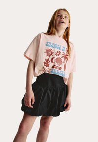 Un t-shirt rose à manches courtes avec des graphiques colorés sur le thème de l'océan et du texte. Associé à une jupe noire bouffante arrivée au genou.