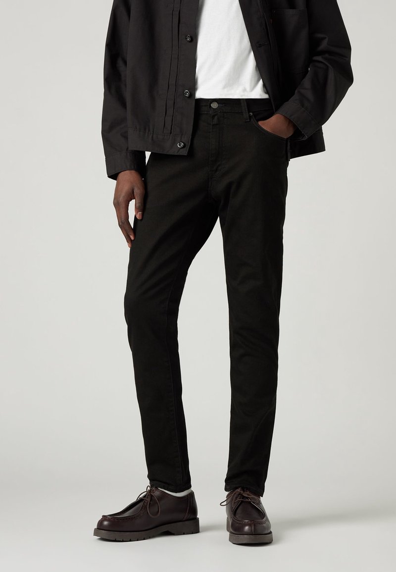 Pantalones de mezclilla negros con ajuste slim, que cuentan con bolsillos frontales y un botón en la cinturilla. Combinados con una chaqueta negra y zapatos marrones.