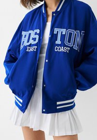 Person som bär en blå Boston varsity-jacka med vit text och en vit plisserad kjol mot en enfärgad bakgrund.