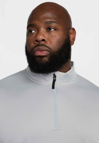 Világosszürke, negyedzáras pulóver, texturált anyagból, fekete cipzárral és álló gallérral. Közeli kép a ruhadarab nyakáról és mellrészéről.