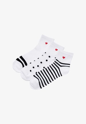 Trzy białe skarpetki do kostki z czarnymi paskami, czarnymi kropkami i czarnymi naszywkami na palcach, każda z małym czerwonym sercem przy mankiecie.