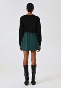 Maglione nero a maglia con dettagli a taglio, abbinato a una gonna verde sopra il ginocchio e stivali neri lucidi al ginocchio.