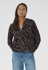 Zwarte blouse met een diepe V-hals, met een golvend rood en wit patroon. Lange, comfortabele mouwen en lichtgewicht stof. Draag met denim jeans.