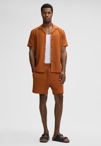 Modello maschile che indossa una camicia a maniche corte button rust colorata e pantaloni corti abbinati, una canottiera bianca e sandali neri, in piedi davanti a uno sfondo neutro.