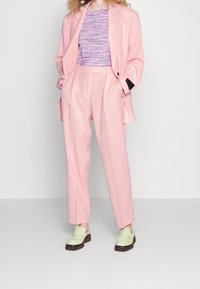 Blazer rose cintré sur un haut à rayures multicolores, associé à un pantalon rose et des mocassins verts avec des semelles brunes.