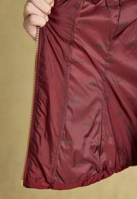 Maroon zippered jacket interior with shiny gold zipper, featuring a smooth, lightweight fabric and subtle black logo pattern.