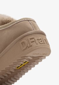 Gros plan sur le talon d'une chaussure en suede beige avec le nom de la marque "D.Franklin" embossé sur la semelle en caoutchouc et un logo jaune sur la bande de roulement.