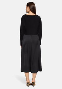 Femme aux longs cheveux bruns portant un pull court noir, une jupe midi noire et des sandales à talons hauts noires, vue de dos.