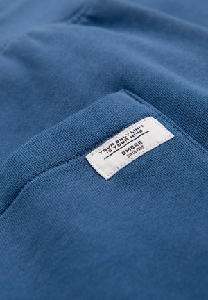 Tissu bleu avec une étiquette rectangulaire blanche cousue, affichant le texte « VOTRE SEULE LIMITE EST VOTRE ESPRIT » et « OMBRE depuis 1995 » sur le bord d'une poche.