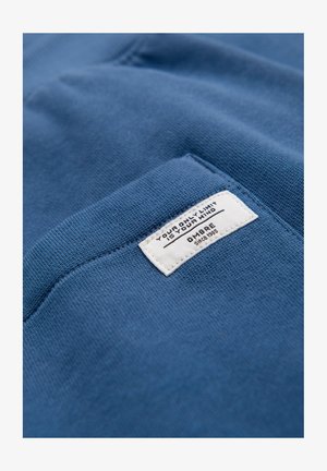 Tissu bleu avec une étiquette rectangulaire blanche cousue, affichant le texte « VOTRE SEULE LIMITE EST VOTRE ESPRIT » et « OMBRE depuis 1995 » sur le bord d'une poche.