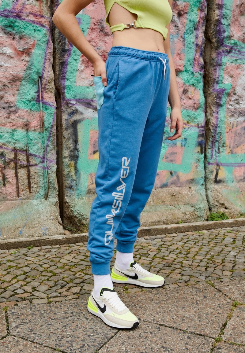 Light blue sweatpants with elastic waistband, side pockets, and "Quiksilver" logo. Paired with neon yellow and black sneakers on cobblestone.
