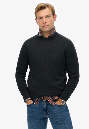 Donkergrijze gebreide trui met een ronde halslijn, gelaagd over een rood, groen en marineblauwe geruite blouse, gedragen met blauwe jeans en een bruine horloge.