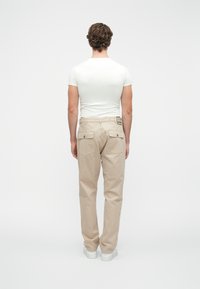 Bežīgas bikses ar taisnu griezumu, divas aizmugurējās kabatas ar pogu aizdari un teksturētu audumu. Valkā ar pieguļošu baltu T-kreklu.