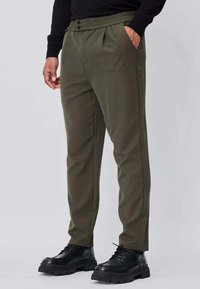 Pantalon vert olive avec une ceinture élastique, des plis et des poches latérales, associé à des chaussures noires épaisses à lacets. Texture de tissu mate.