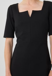 Robe noire ajustée avec des manches courtes, décolleté carré et une légère encolure en V. Le tissu semble lisse avec des coutures minimales et une coupe ajustée.