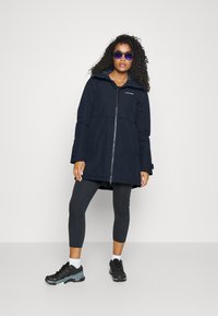 Veste imperméable bleu marine avec capuche, fermeture éclair avant intégrale et deux poches latérales ; associée à des leggings noirs et des baskets sombres.