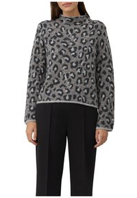 Vrouw in een grijs en zwart panterprint trui met lange mouwen en zwarte broek, staand tegen een witte achtergrond.