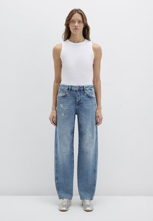 Hellblaue, abgenutzte Jeans mit weitem Bein und hoher Taille, kombiniert mit einem engen weißen gerippten Tanktop und silberfarbenen Turnschuhen.