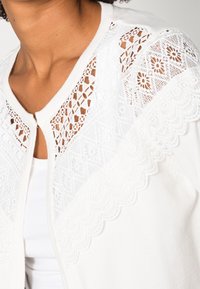 Bílý kardigan s jemnými krajkovými detaily na ramenou, zakončený vroubkovanými lemy a texturovaným vzorem. Přední zapínání na zip.