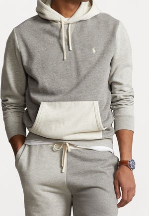 Uomo che indossa una felpa con cappuccio bicolore grigio chiaro e pantaloni della tuta abbinati con le mani nelle tasche e un orologio da polso con cinturino in pelle marrone visibile.