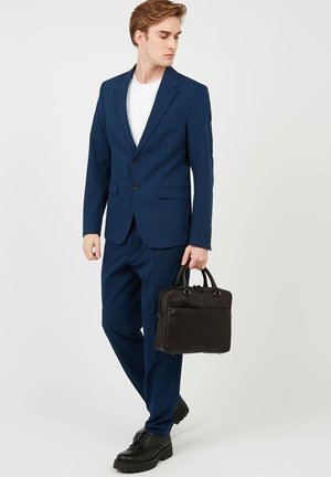 Costume bleu marine avec une veste cintrée et un pantalon assorti. Porté avec un t-shirt blanc. Tient une mallette en cuir noir. Chaussures de ville noires.