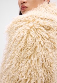 Gros plan sur une personne portant une veste en fausse fourrure bouclée de couleur crème, montrant une partie du visage et de l'épaule.