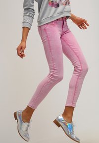 Henkilö, joka on pukeutunut vaaleanpunaisiin kapeisiin farkkuihin ja metallinhohtosiin nauhakenkiin, hyppää yksinkertaista vaaleaa taustaa vasten.