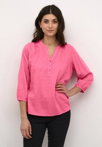 Blouse en lin rose avec un col en V et une patte de boutonnage à trois boutons. Dispose de manches trois-quarts avec des poignets élastiques et d'une coupe décontractée.
