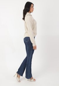 Beige gerippter Pullover mit Kragen, kombiniert mit dunkelblauen Glockenjeans und hellen Schuhen mit einem gesteppten Muster.