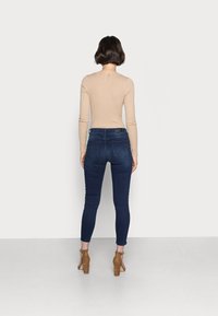 Nainen, jolla on lyhyet ruskeat hiukset, pukeutuneena beigeen ribbitoppiin, tummansinisiiin skinny-farkkuihin ja ruskeisiin korkokenkiin, seisoo tasaisen seinän against.