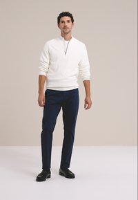 Weißer Pullover mit Reißverschluss am Kragen, marineblaue Hose und schwarze Schuhe. Der Pullover hat gerippte Bündchen und eine glatte Textur, während die Hose maßgeschneidert ist.