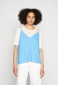 Top azul sem mangas com alças ajustáveis sobre uma camisa de manga curta branca. Textura suave e corte solto, com um design em V e bainha reta.