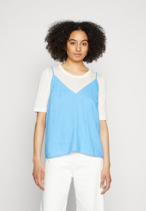Top azul sem mangas com alças ajustáveis sobre uma camisa de manga curta branca. Textura suave e corte solto, com um design em V e bainha reta.