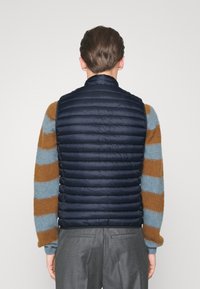 Marineblå dunvest med horisontalt quiltet design. Letvægts, med lynlåslukning foran. Båret over en stribet, textured sweater i dæmpede farver.
