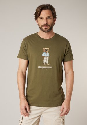 Hombre con camiseta verde oliva con un dibujo de un oso con gafas de sol y ropa informal, combinada con pantalones beige claros.