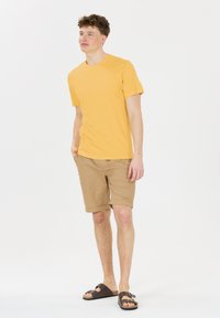 Gul bomull t-shirt, beige shorts och bruna sandaler. T-shirten har en rund halsringning; shortsen är uppvikta i fållen. Enkelt avslappnat outfit.
